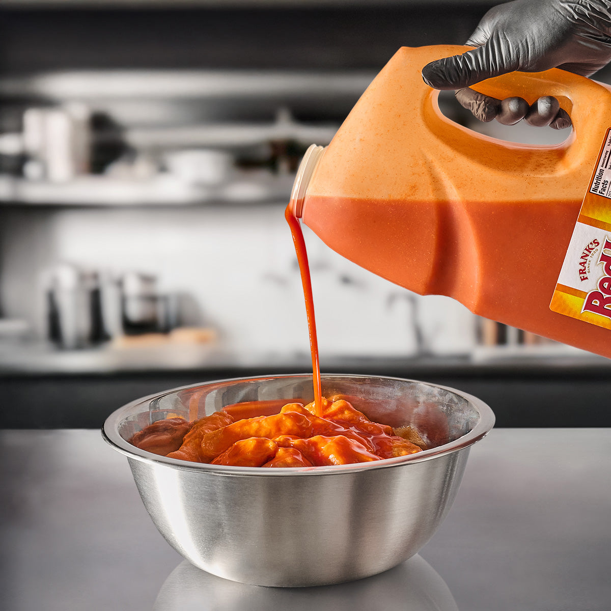 Franks RedHot Sauce being poured onto a bowl with heaping chicken wings on a stainless steel table