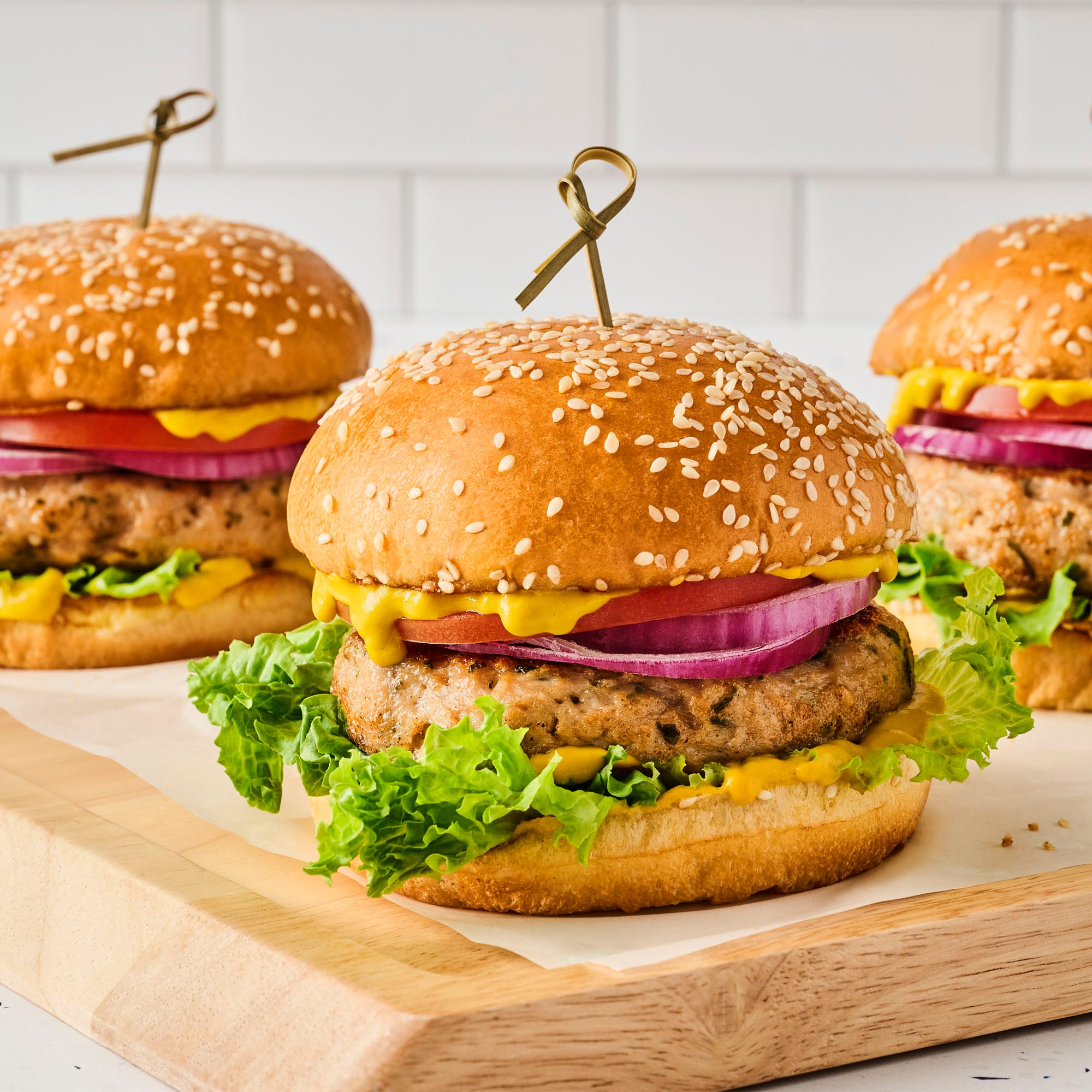 Three burgers on a wooden board with a white tiled wall background