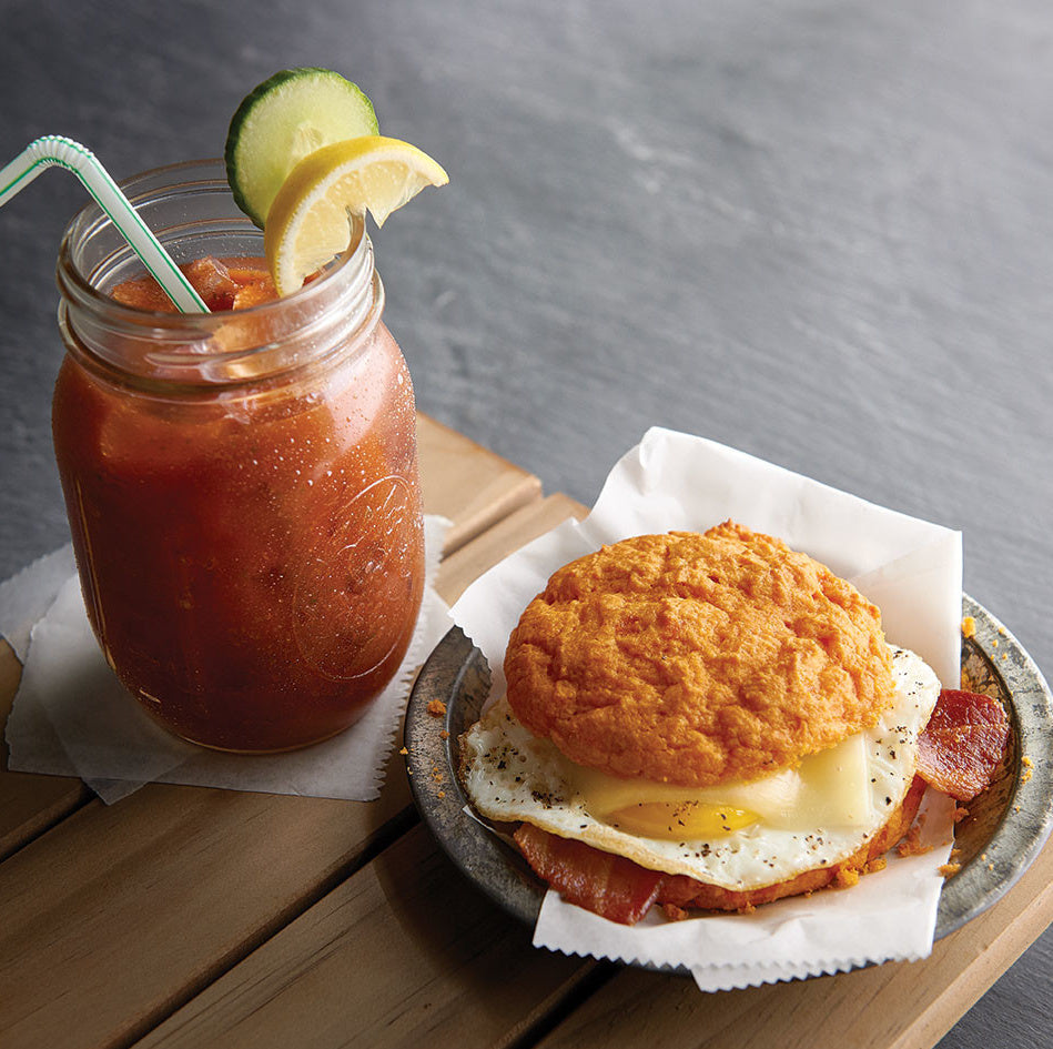Bloody Mary with a lemon wedge and straw, alongside a breakfast sandwich on a wooden table.