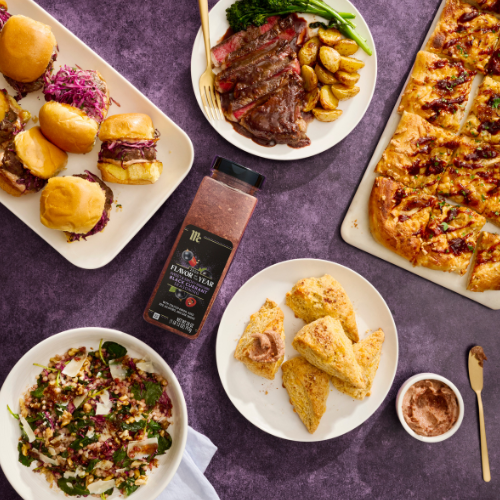 Assorted food items including burgers, steak, bread, and salad on a purple surface with a bottle of black currant seasoning.