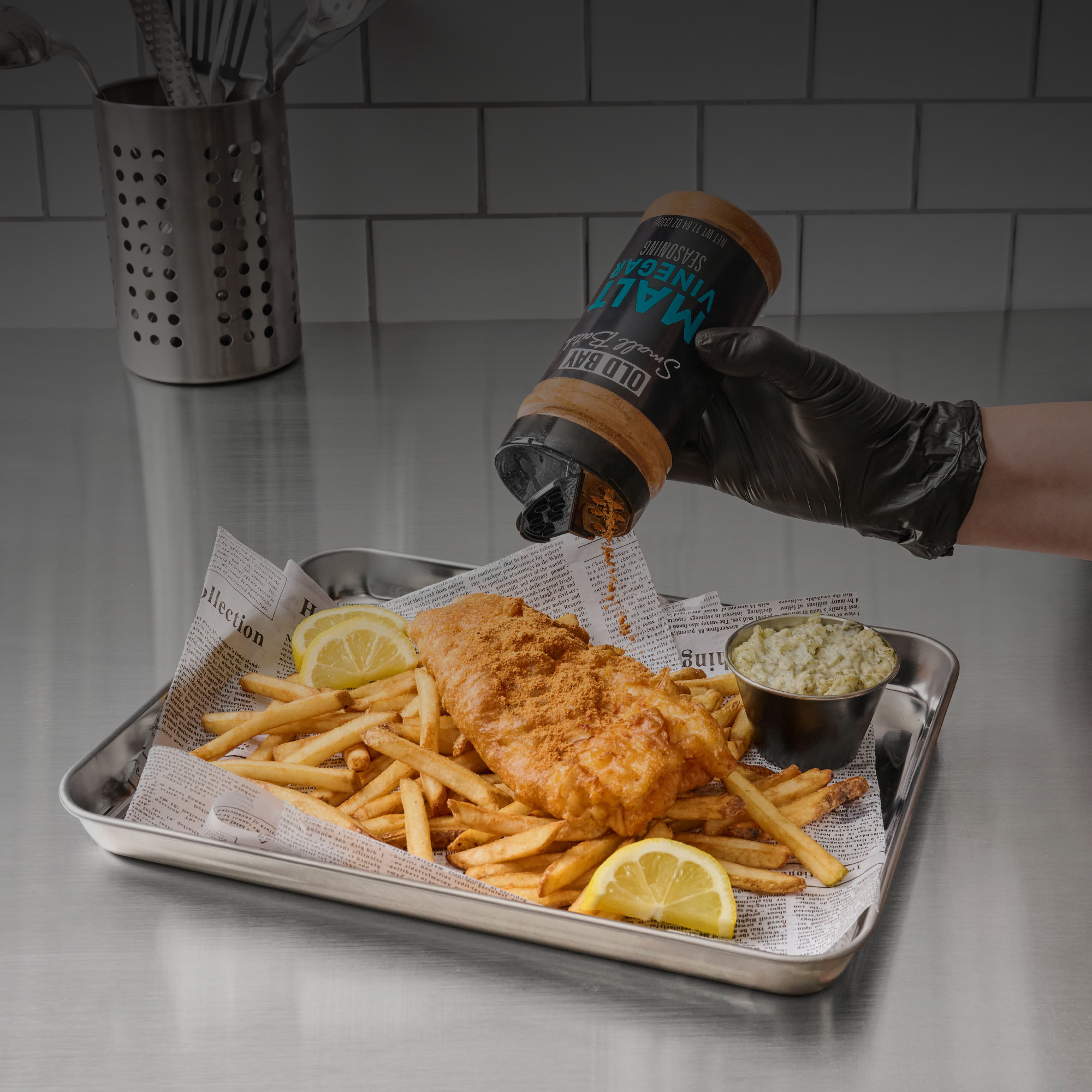 Person seasoning fish and chips with a bottle of malt vinegar on a metal tray.