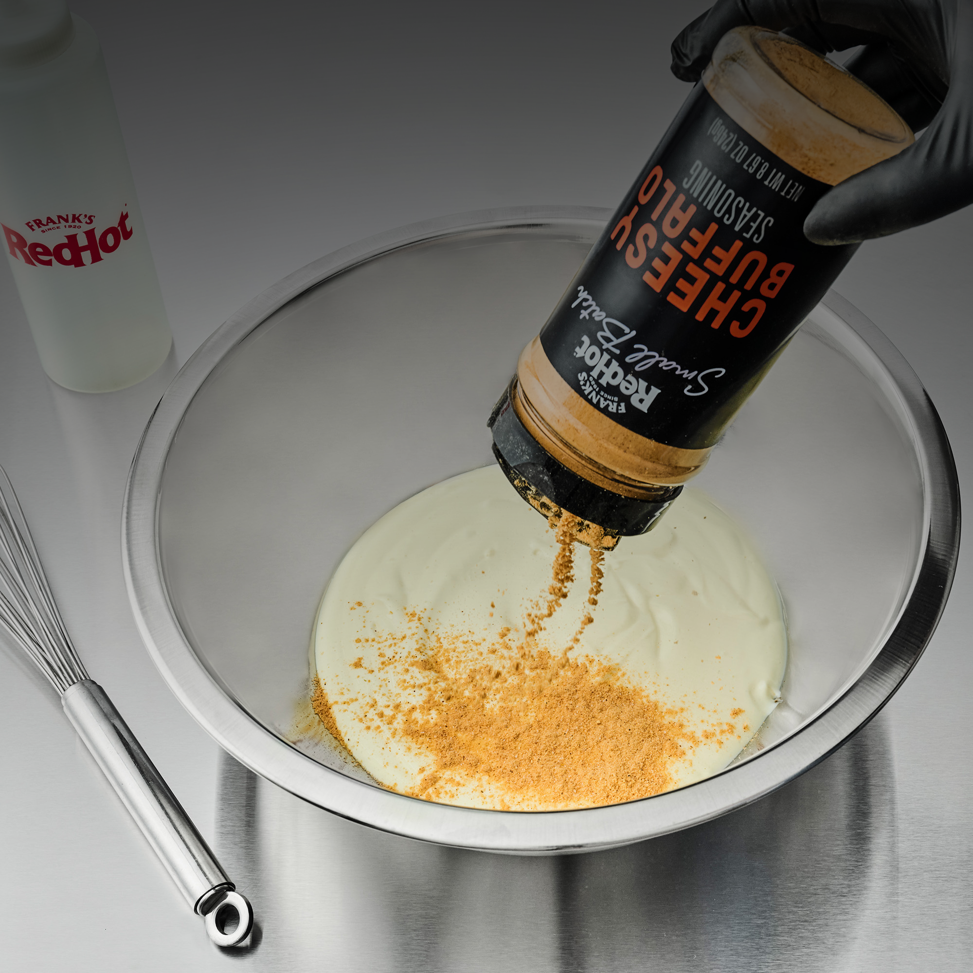 Hand sprinkling Buffalo Cheese sauce into a bowl with a whisk and bottle of Frank's RedHot in the background.