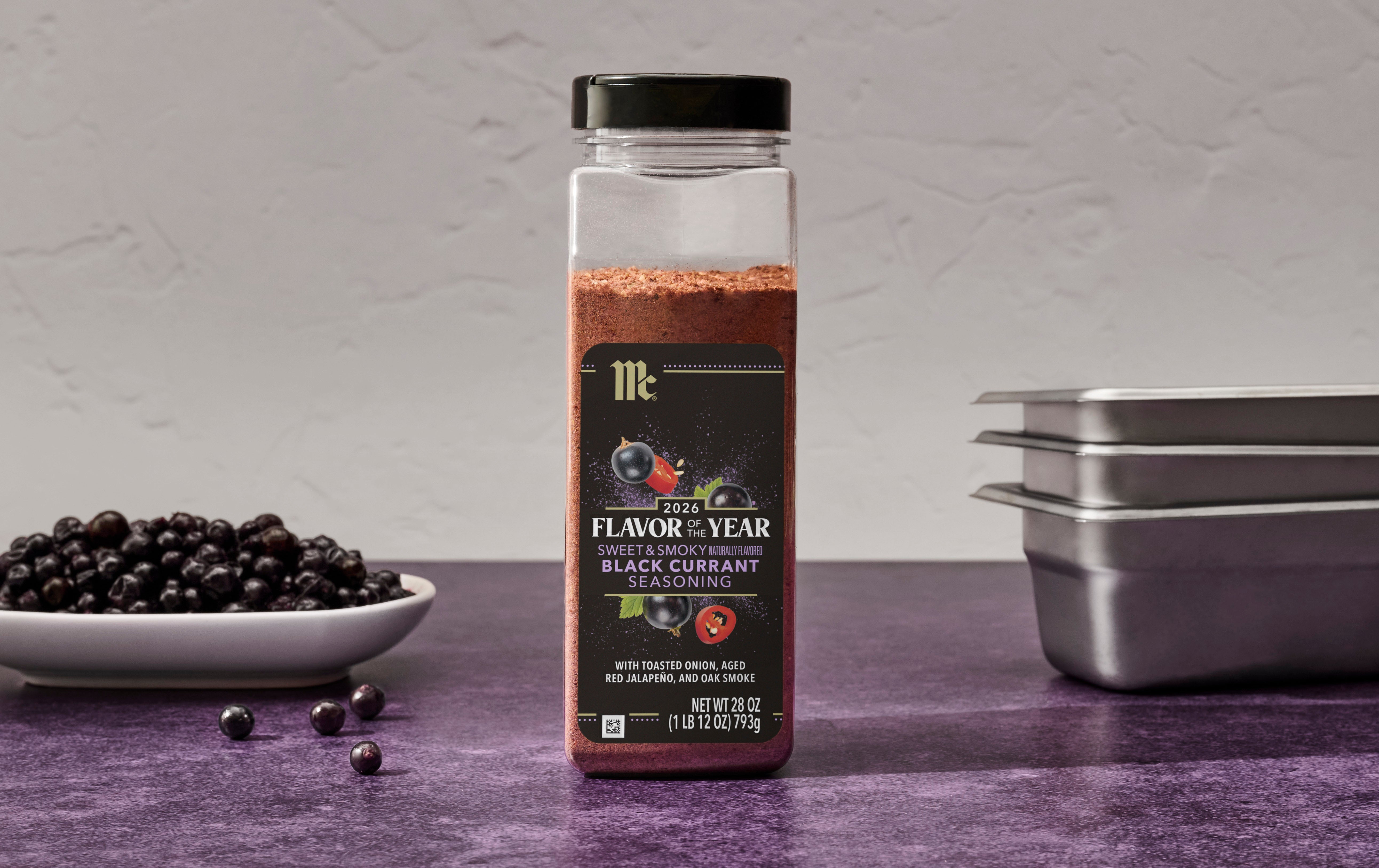Bottle of McCormick Black Currant seasoning on a purple surface with black currants and metal pans in the background.