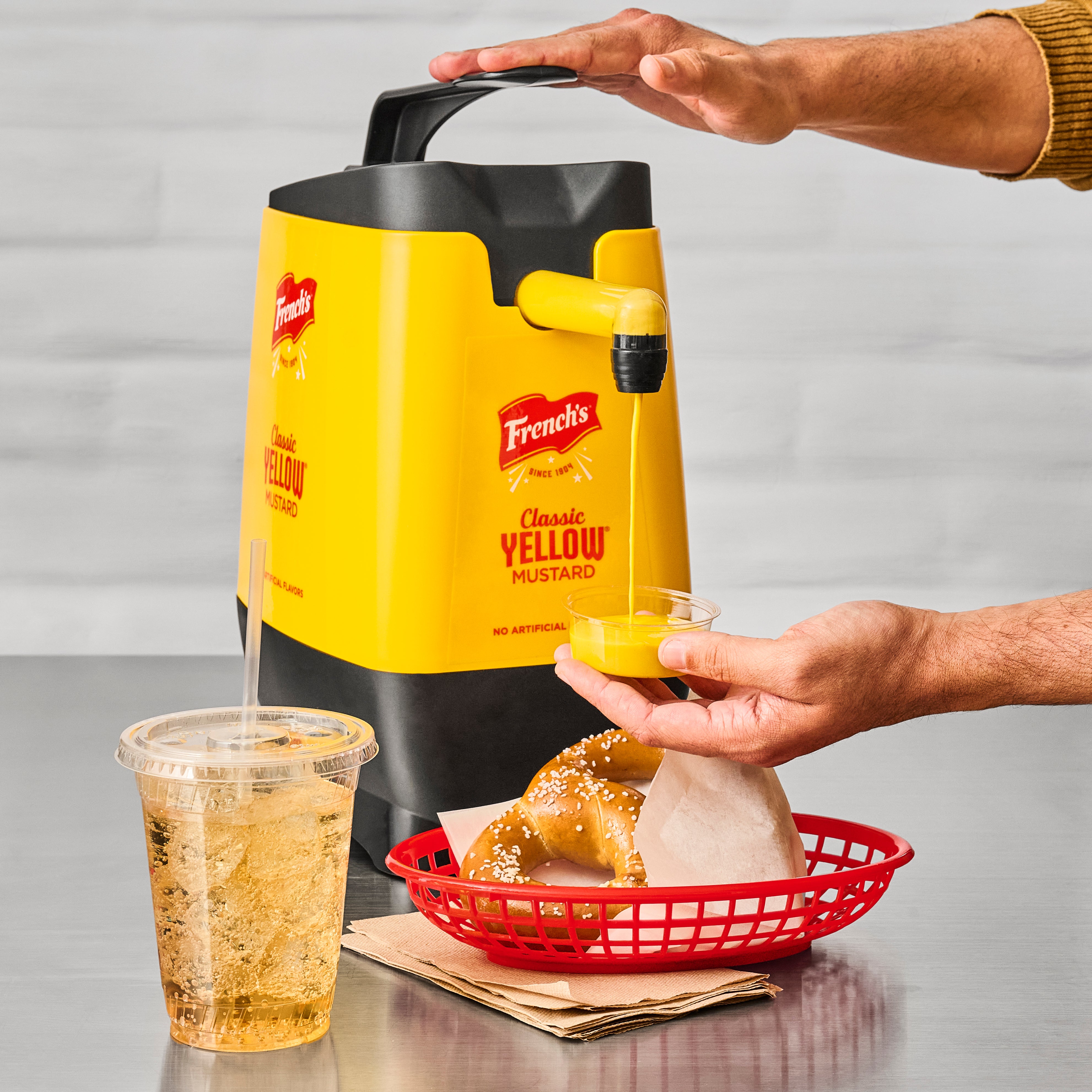 Person using a mustard dispenser to apply mustard on a pretzel with a drink and basket in the background.